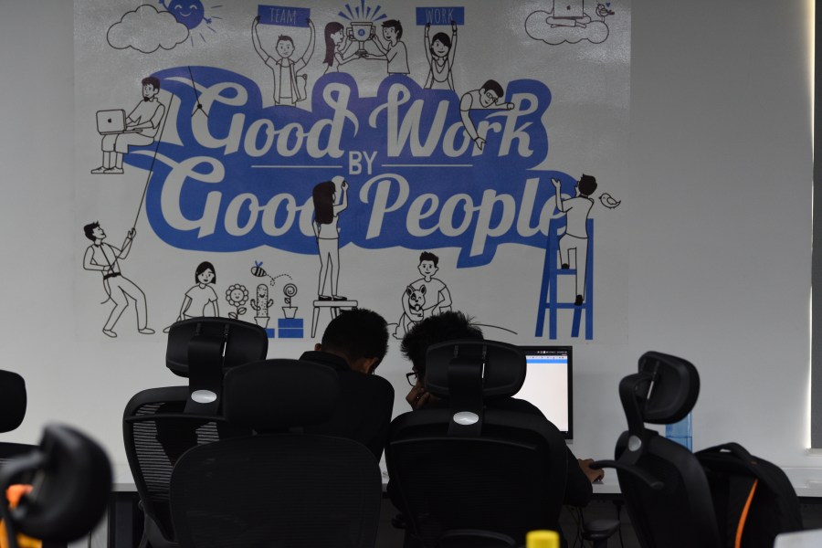 Two people are silhouetted at their desks, with a poster on the wall in front of them that says “Good Work by Good People”