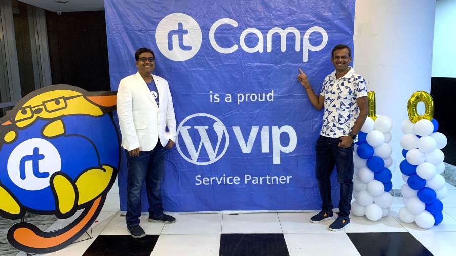 At WordCamp Kolkata, CEO of rtCamp Rahul Bansal wears a white blazer and poses next to VIP Anand Natarajan in front of a blue banner celebrating rtCamp as a featured partner of WordPress VIP