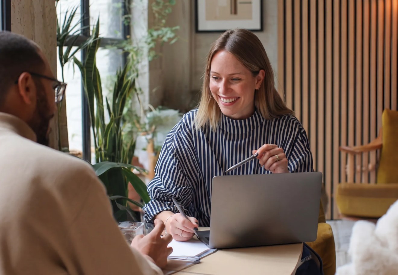 Woman smiling with laptop at a table, discussing content marketing for business growth with a man in a bright, modern office space.