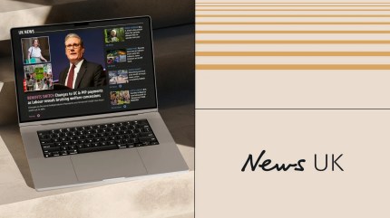 Laptop displaying the News UK homepage with breaking headlines, next to the News UK logo on a beige and gold-toned branded background.
