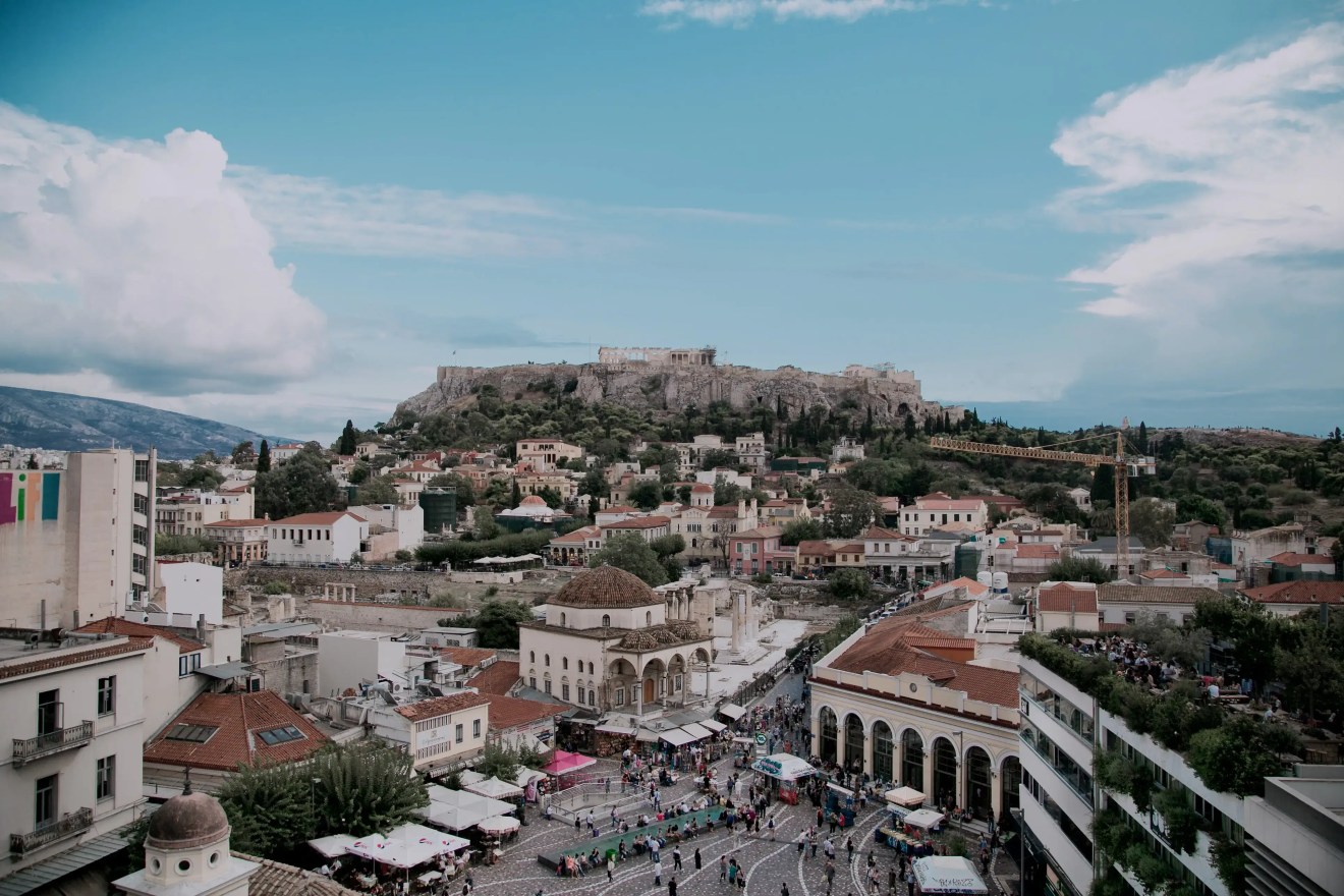 The Acropolis of Athens rising above a busy city square with historical buildings and crowds of people.