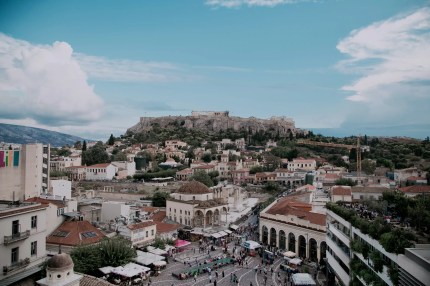 The Acropolis of Athens rising above a busy city square with historical buildings and crowds of people.