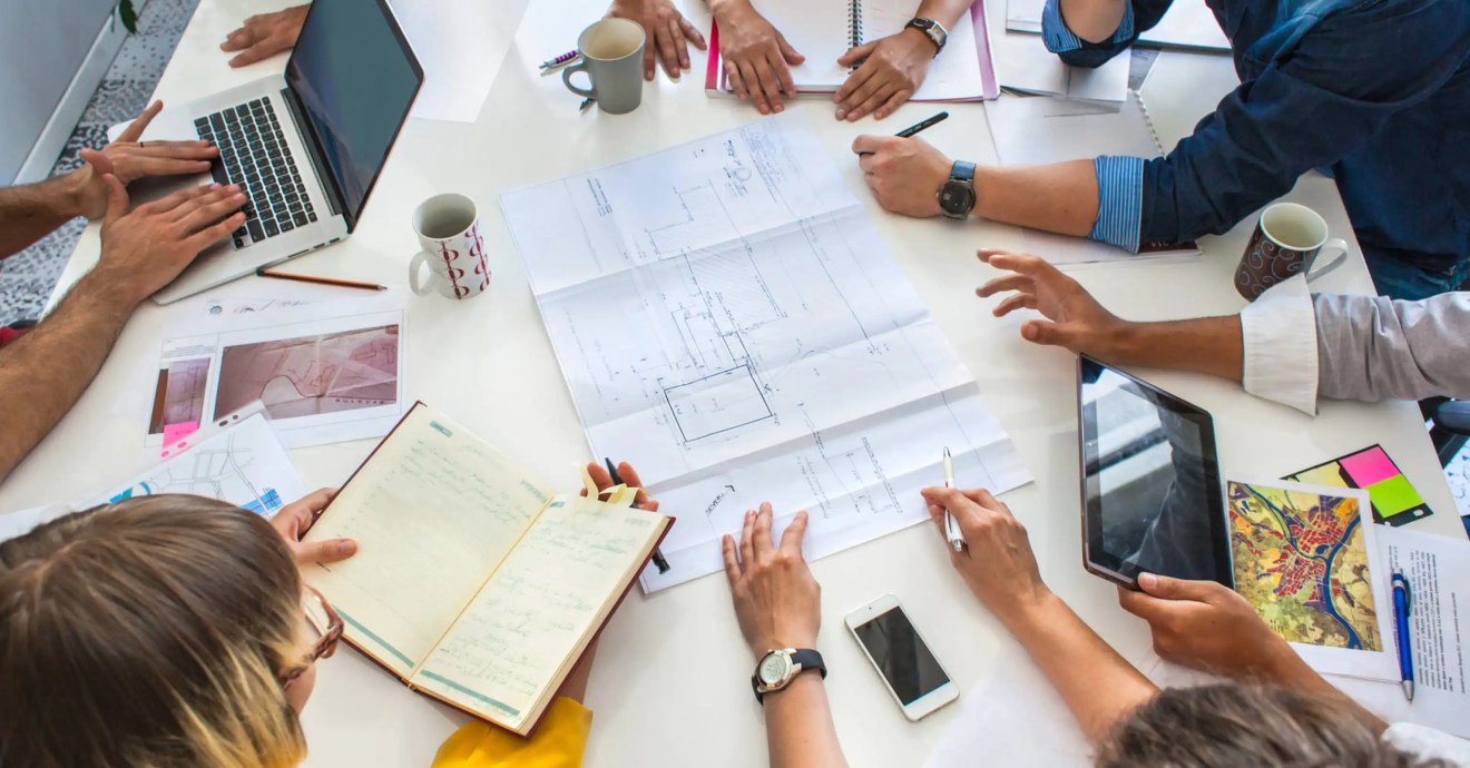 A group of people circled around a plan, each person has a combination of pens, notebooks, laptops, tables, etc for collaborating.