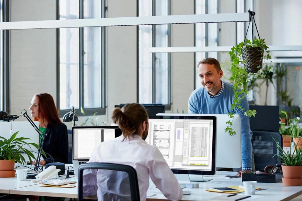 Two men discuss editorial workflow at a digital solution desk with computers; nearby, a woman focuses on content tasks.