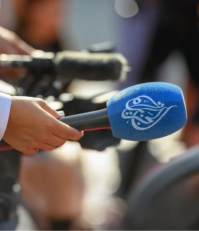 Close-up of a hand holding a blue foam-covered microphone with the Al Jazeera logo, aimed toward an unseen speaker. A camera and recording equipment are visible in the blurred background.