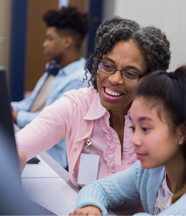 Teacher assisting a student at a computer in a classroom while other students work in the background.