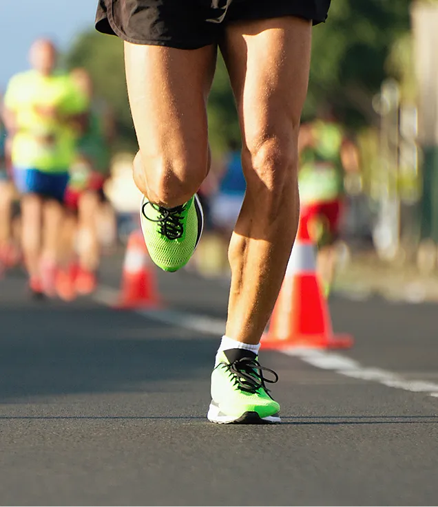 Close-up of a runner's legs wearing bright green running shoes during a road race, with traffic cones and other runners blurred in the background.
