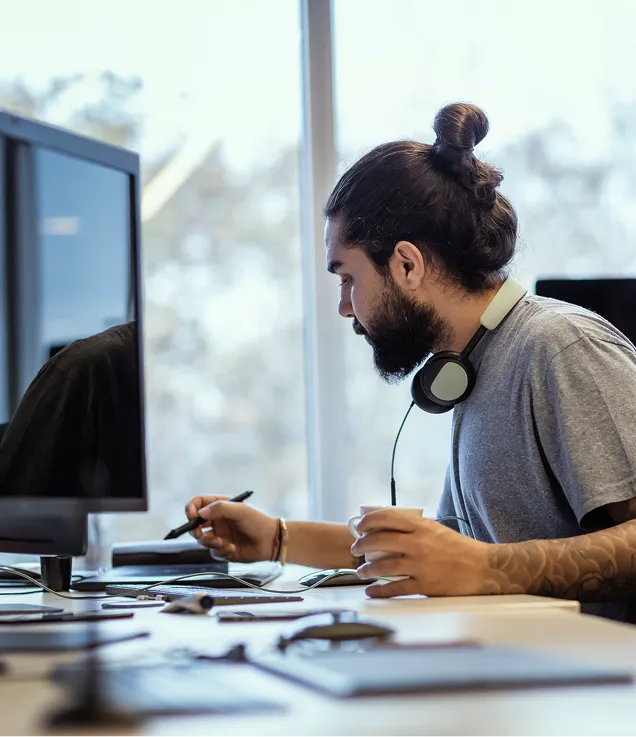 A professional working at a desk with a pen tablet, computer monitor, headphones around their neck, and a coffee cup beside them.