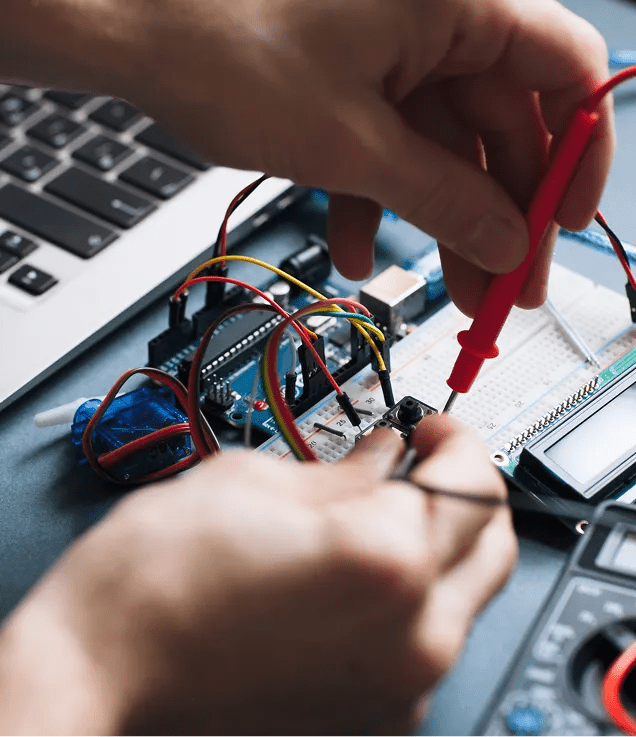 Close-up of hands using multimeter probes to test an electronic circuit connected to a breadboard, microcontroller, and laptop.