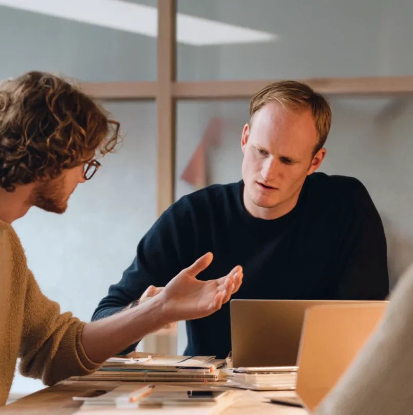 Two colleagues discussing work across a table with laptops, notebooks, and documents.