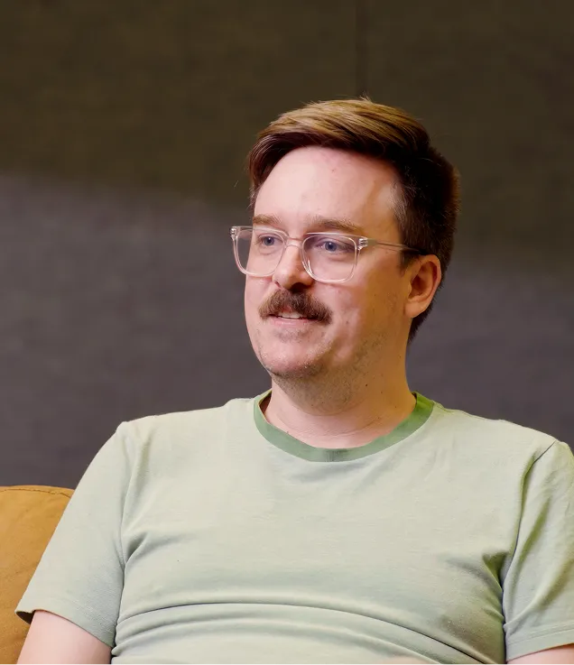 Man with glasses and a mustache wears a light green shirt, sitting on a tan couch against a gray wall.