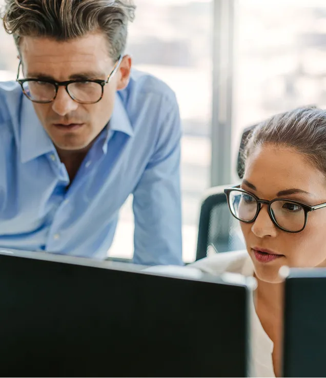 Two people wearing glasses looking at computer monitors in an office. One person is seated while the other leans over to review the screen, suggesting collaboration on a task or project.
