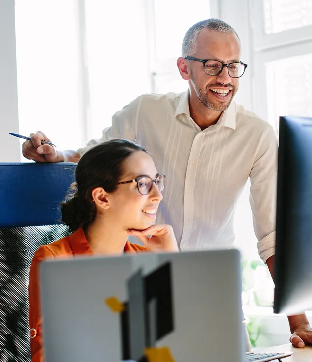 A professional standing beside a seated colleague as they review content together on a desktop computer in a bright office.