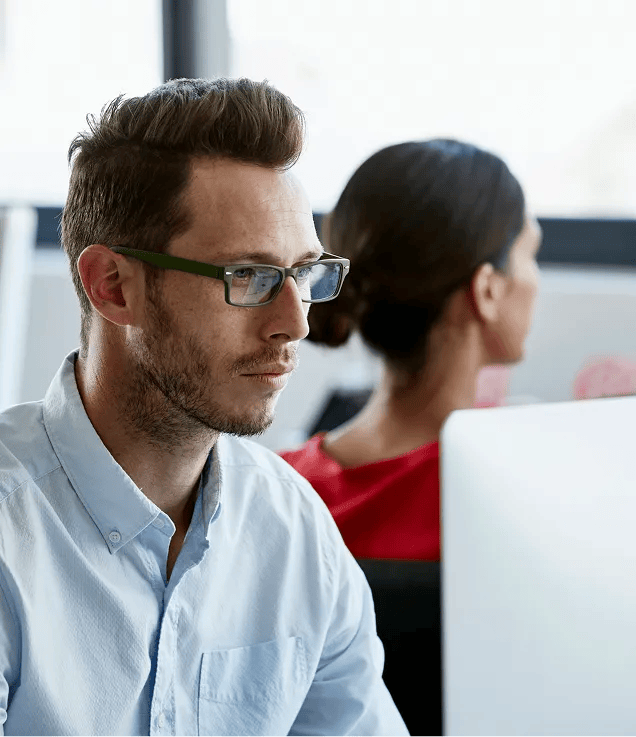 Man with glasses working at computer; woman in red shirt sits blurred in the background.