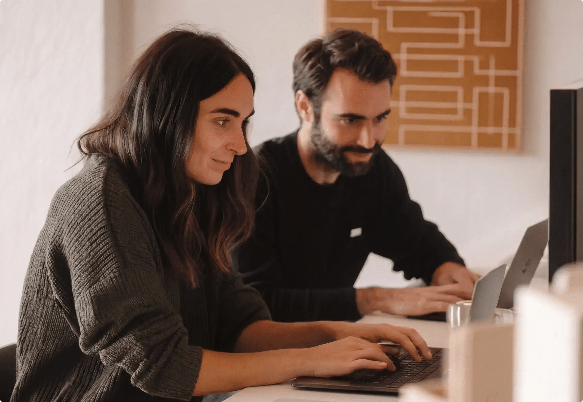 Two people sit at a desk, working on laptops, focused and smiling in a cozy office setting—illustrating the collaborative spirit that flourishes when using WordPress VIP for engineering and IT teams.