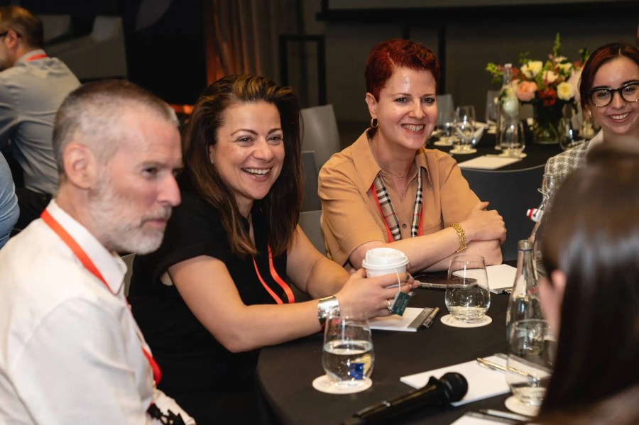 Four people sit at a round table, smiling and talking at the Innovation Exchange event, with drinks and notepads in front of them.