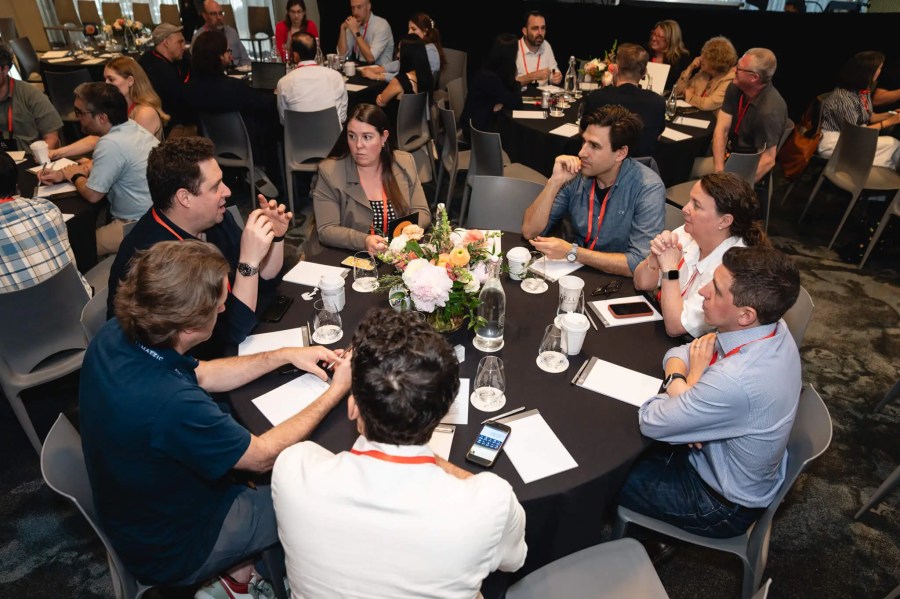 People sit around a round table with papers at the Innovation Exchange event, engaged in discussion, with flowers in the center.