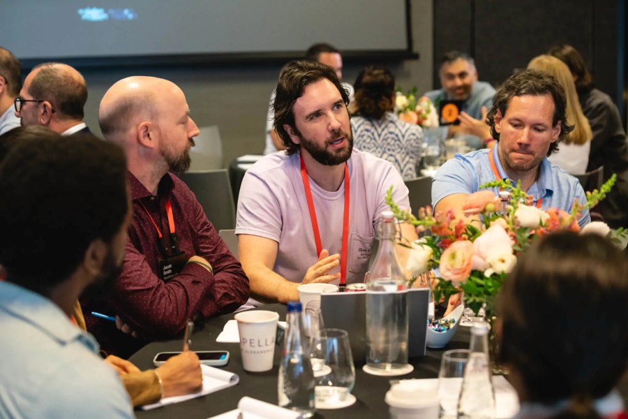 Four men talk at a round table with flowers, water glasses, and papers during a conference.