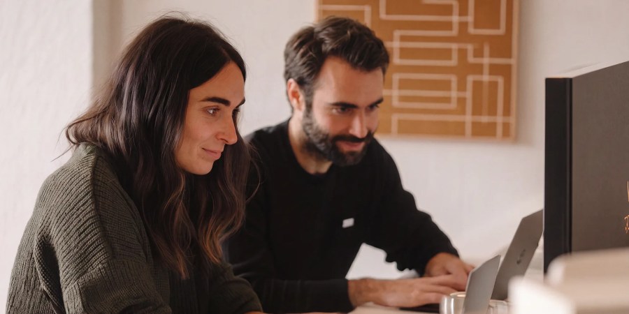 Two people sit side by side at a desk, working on laptops and looking at their screens, smiling slightly as they collaborate with an AI content analytics assistant.