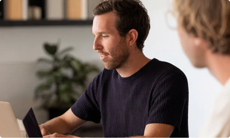Two people sit at a table working on laptops, with shelves and a plant in the background—collaborating seamlessly as they explore insights provided by a content analytics AI assistant.