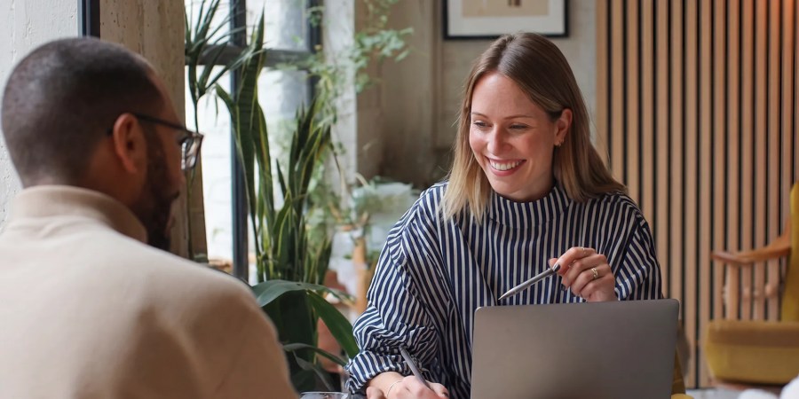 Two people sit at a table with a laptop, having a friendly conversation in a modern office setting as they explore the powerful features of AI content analytics.