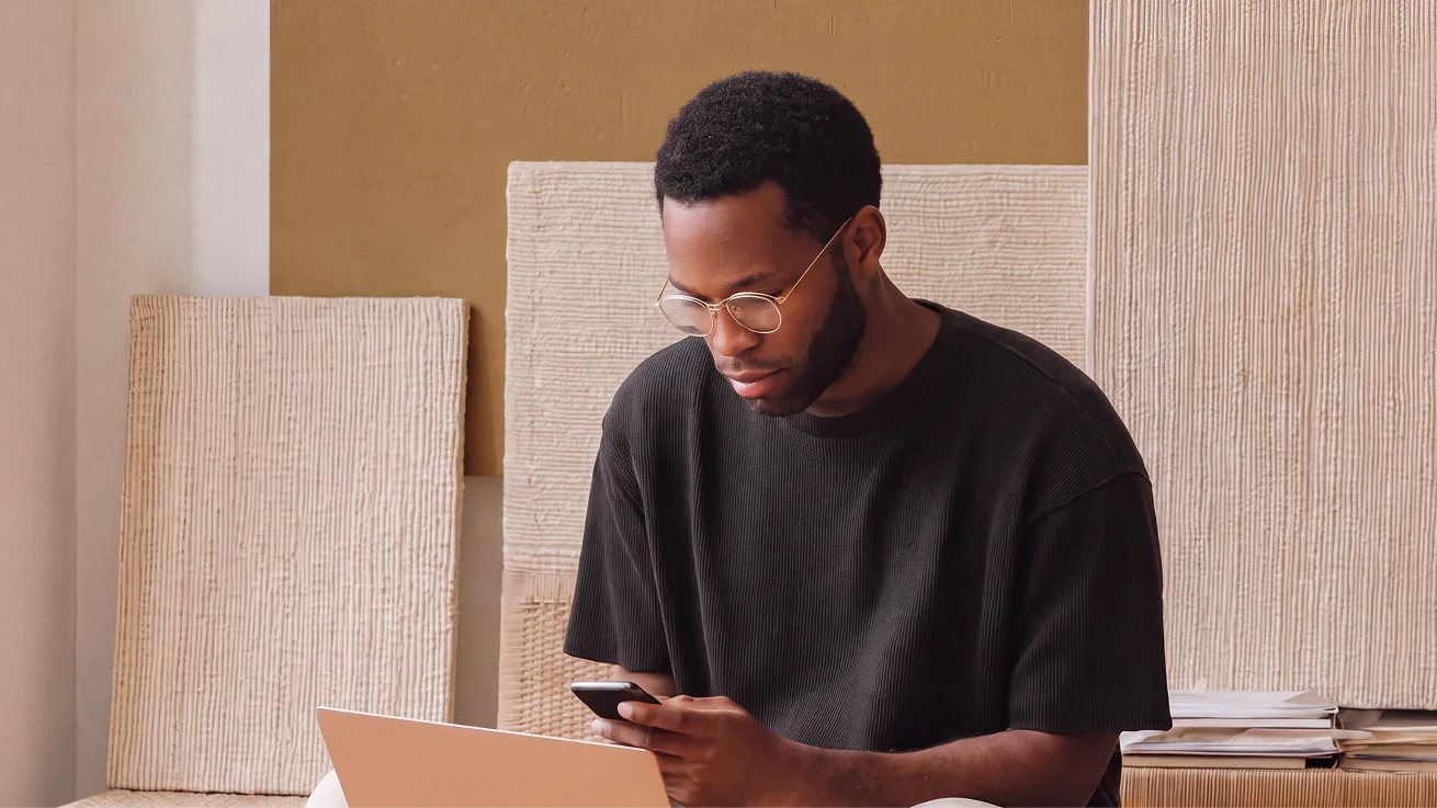 Man wearing glasses sits indoors, looking at his phone and laptop, utilizing a content analytics AI assistant, with beige textured panels behind him.