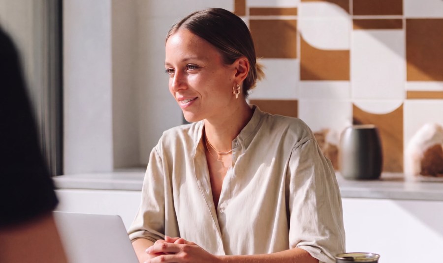 Woman smiling and sitting at a table with a laptop in a bright room with geometric wall tiles.