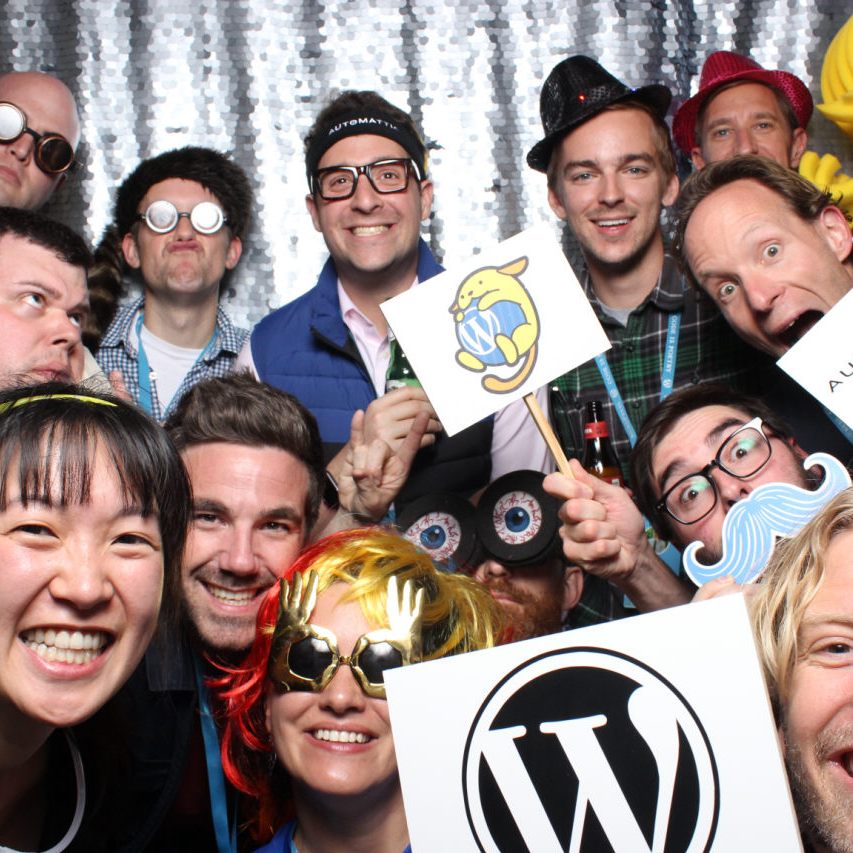 A group of people wearing funny hats and accessories, and holding up WordPress and Automattic signs in a photo booth at a company meetup in Park City.